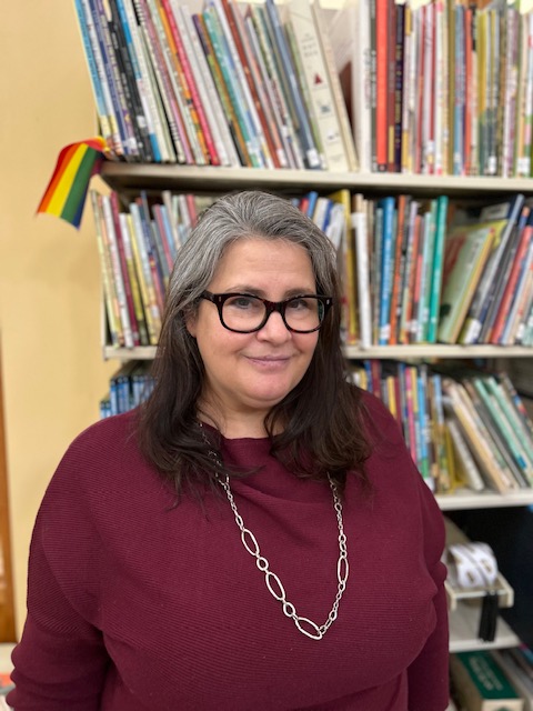 Eileen is smiling in front of a full bookshelf. She is wearing glasses and her gray and brown hair is shoulder length. She is wearing a burgundy sweater and a long silver necklace.