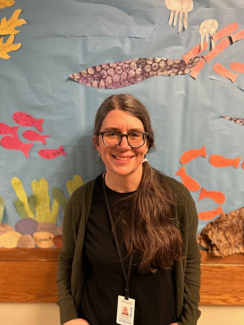 Allison is smiling in front of blue bulletin board with sea creatures and plants. Her long brown hair is tied over her shoulder and she is wearing glasses. She is wearing a black shirt and gray cardigan.