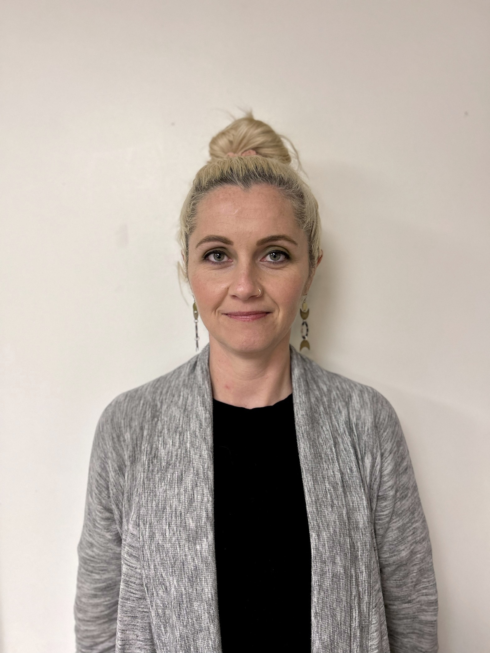 Shannon is in front of a white wall with her blonde hair in a high bun, dangly earrings, a black t-shirt and a gray cardigan.