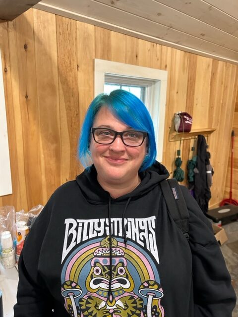 Christina is wearing a black Billy Strings hoodie. She has dark framed glasses and turquoise chin length hair.