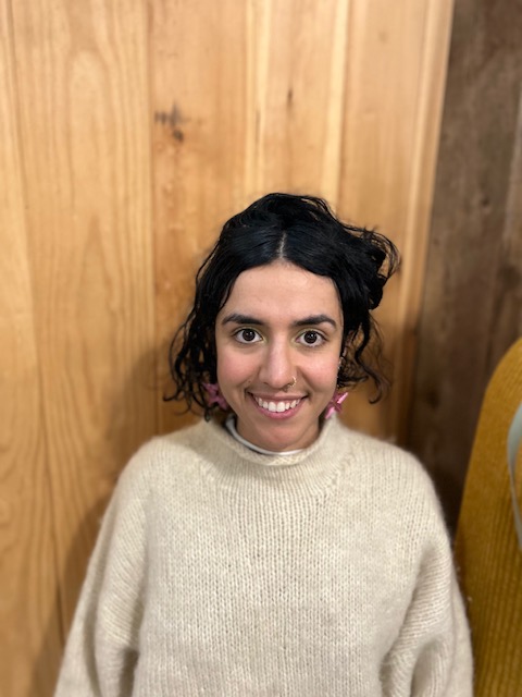 Nina is smiling in front of a wooden wall. Their dark hair is swept back and they are wearing a cream colored sweater.