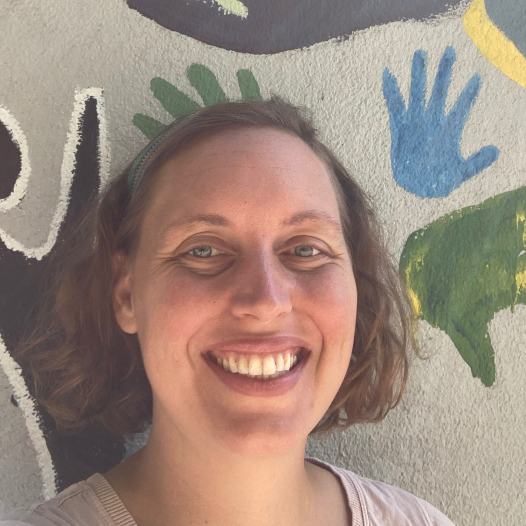 Nicole is smiling in front of an off-white wall. she is wearing a black shirt with a gray zip up over it, and her long, straight, dark hair is down over her shoulders.