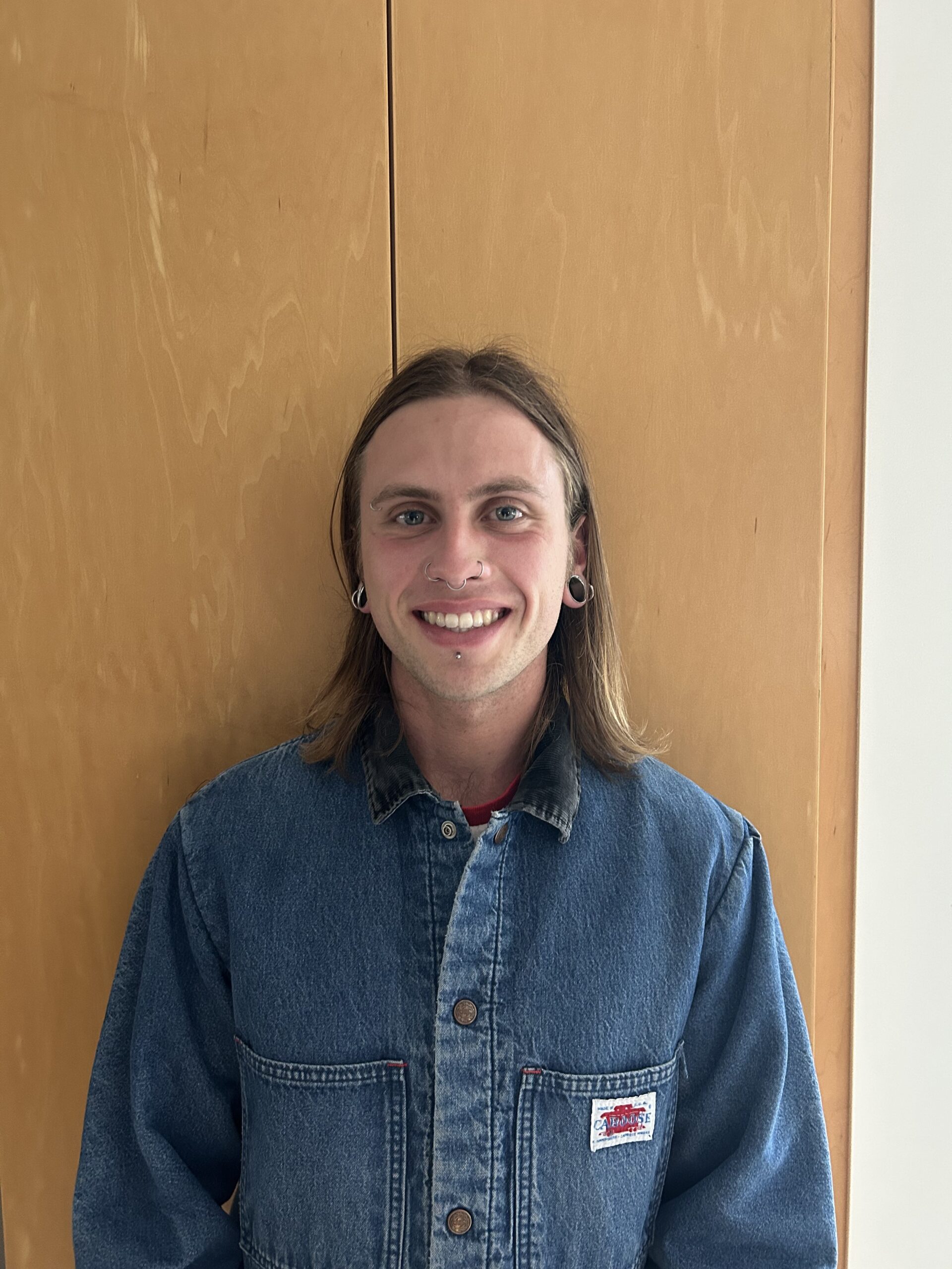 Isaac has straight shoulder length hair, and is smiling in a denim button down.
