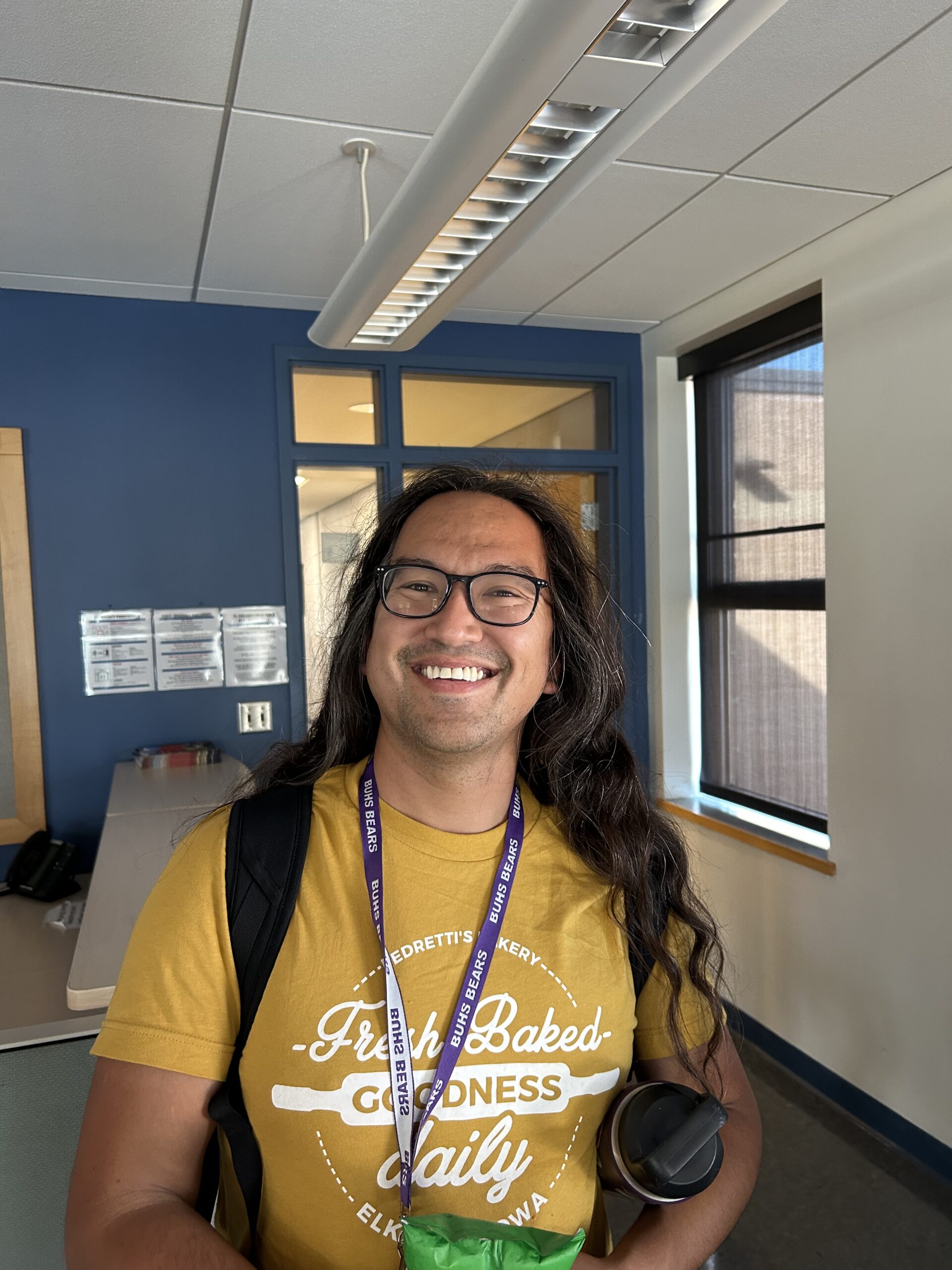 Tyler wears glasses and has long wavy brown hair. They are wearing a yellow tshirt, lanyard and backpack.