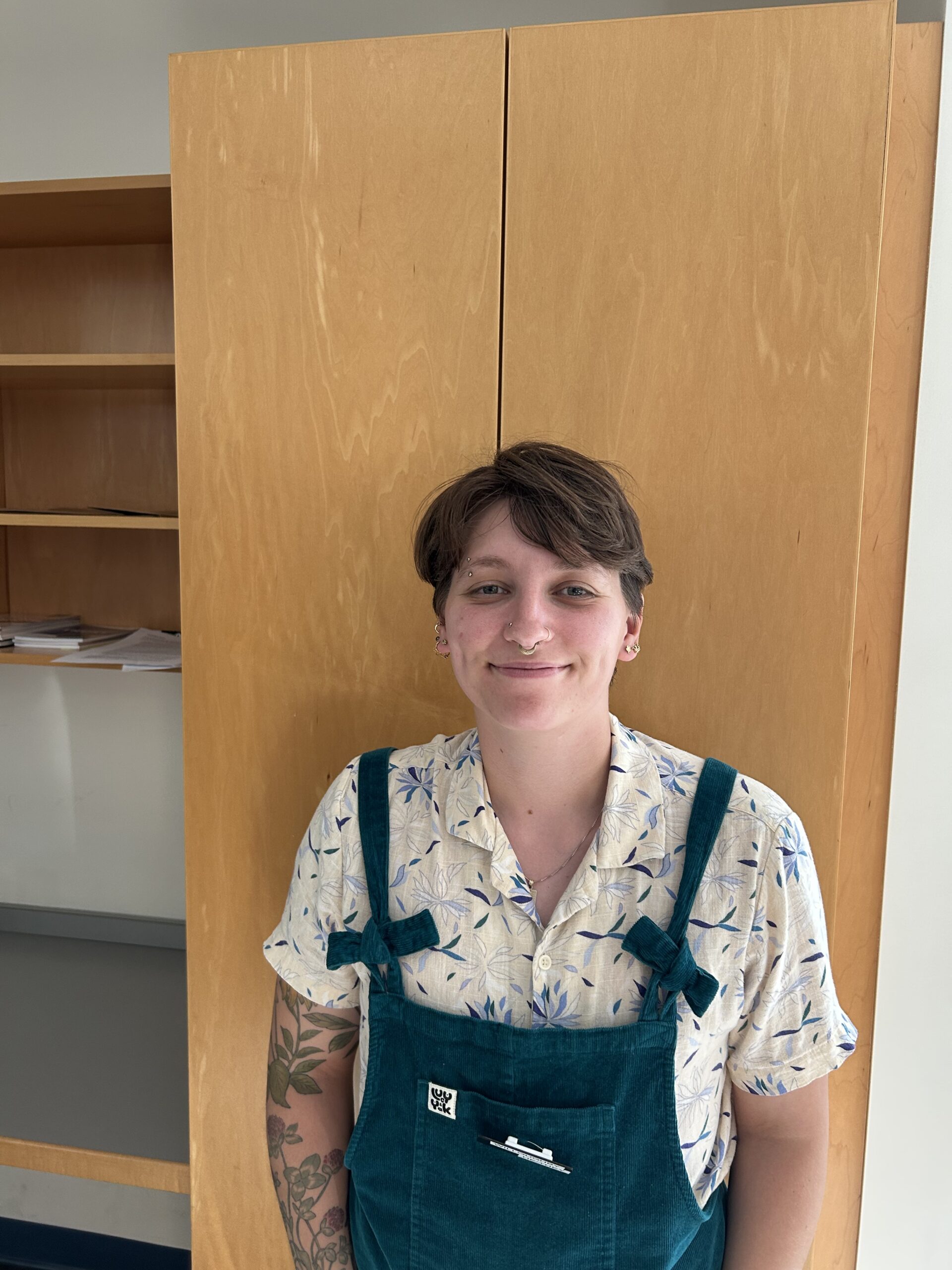 Willoughby has short brown hair and wears green overalls and a white printed button down.